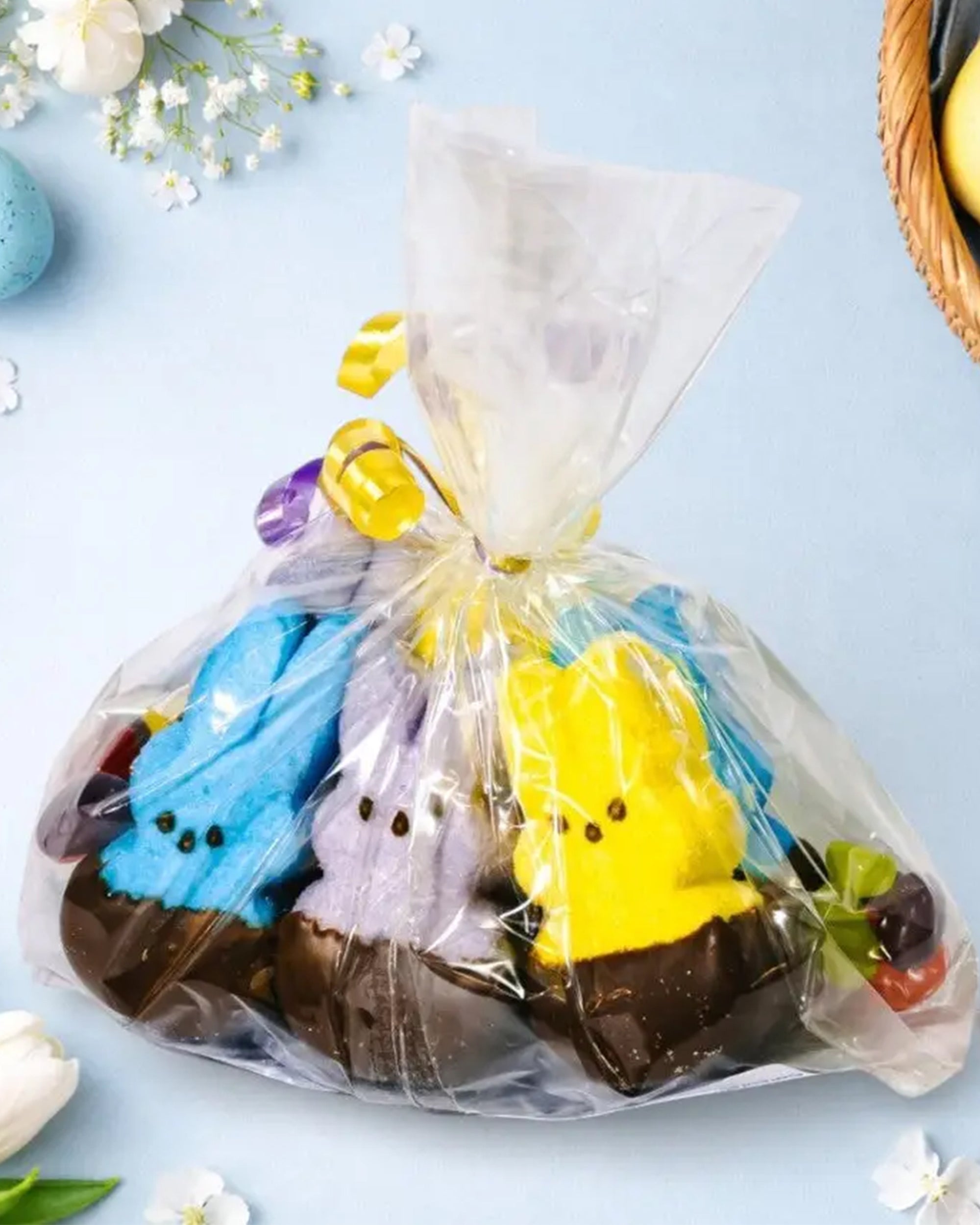 A clear gift bag tied with yellow ribbon holds Faire Easter Dipped Peeps—blue, purple, and yellow bunnies dipped in milk chocolate—surrounded by small candies on a pastel blue surface near flowers and an Easter basket.