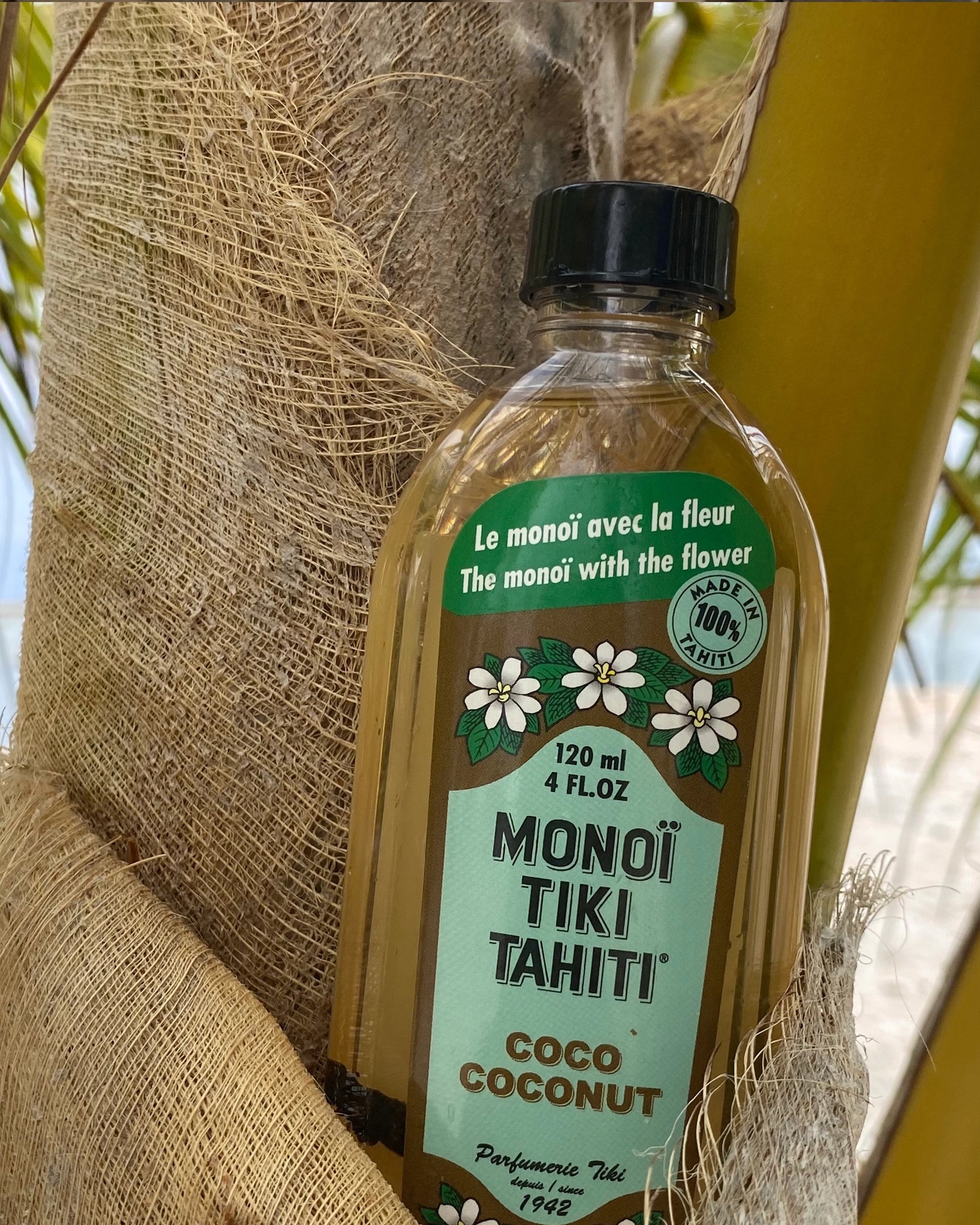 A bottle of Faire Monoi Coco Coconut Oil with a green label and white Tiare flower illustrations rests against a palm tree, set on a sandy beach with lush palm fronds in the background.