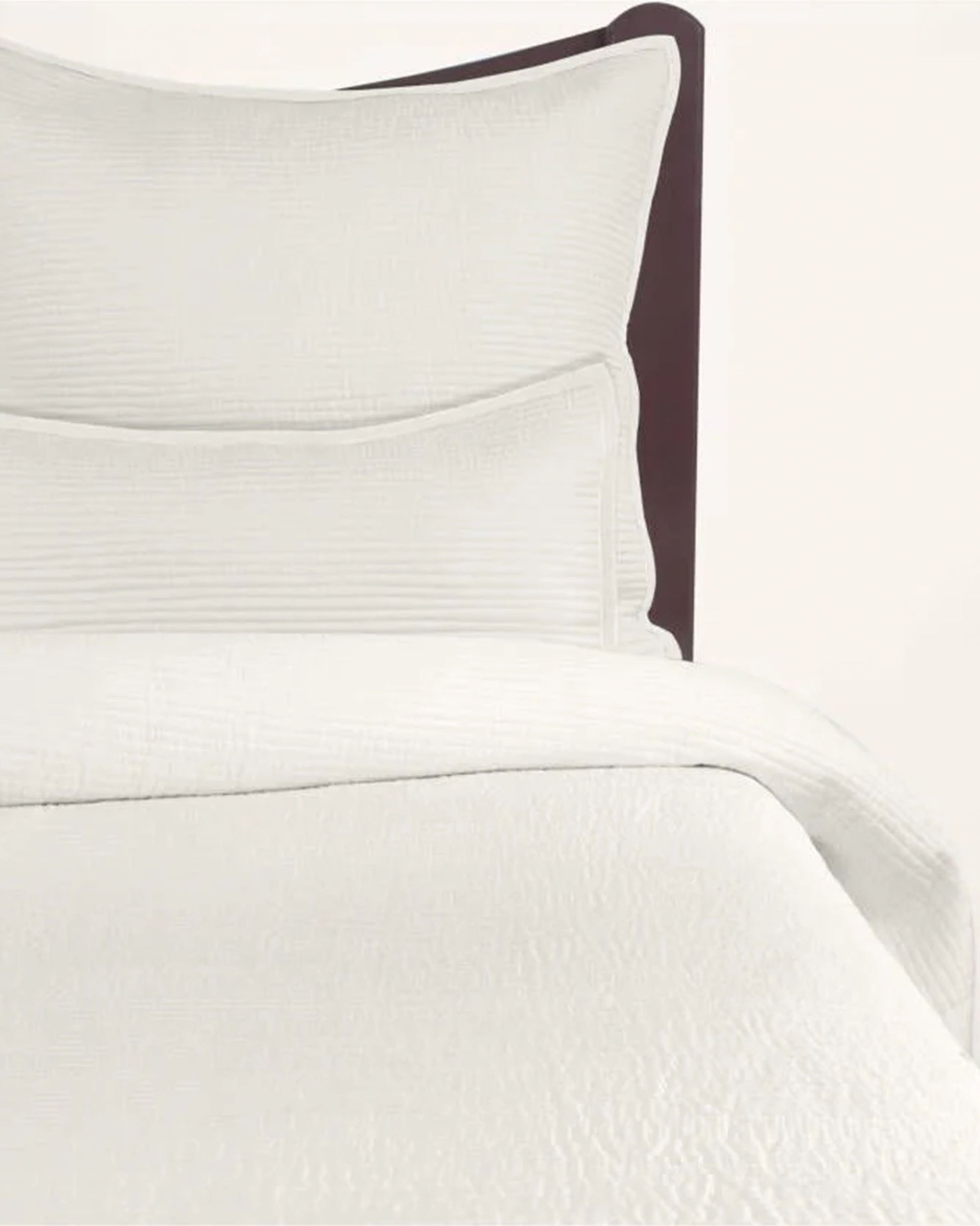 The Cole Quilt Cream King by Avasa Bedding features off-white, textured luxury silk bedding with two matching pillows, displayed on a simple dark brown headboard and a plain light backdrop.