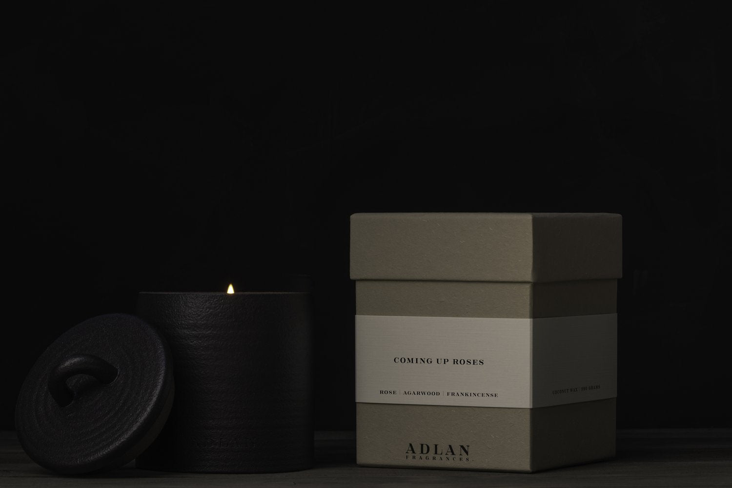 A lit ADLAN Candle with a textured lid sits beside a closed green box labeled “Coming Up Roses,” infused with notes of rose, sandalwood, and rich frankincense. The brand name "ADLAN Fragrances" is elegantly printed on the box, all set against a dark background.