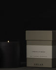 A lit ADLAN Candle with a textured lid sits beside a closed green box labeled “Coming Up Roses,” infused with notes of rose, sandalwood, and rich frankincense. The brand name "ADLAN Fragrances" is elegantly printed on the box, all set against a dark background.