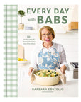 A smiling woman in glasses and a blue floral apron holds a casserole dish in her bright kitchen. She displays "Every Day With Babs" by Random House, your go-to source for 101 delicious, family-approved dinners.