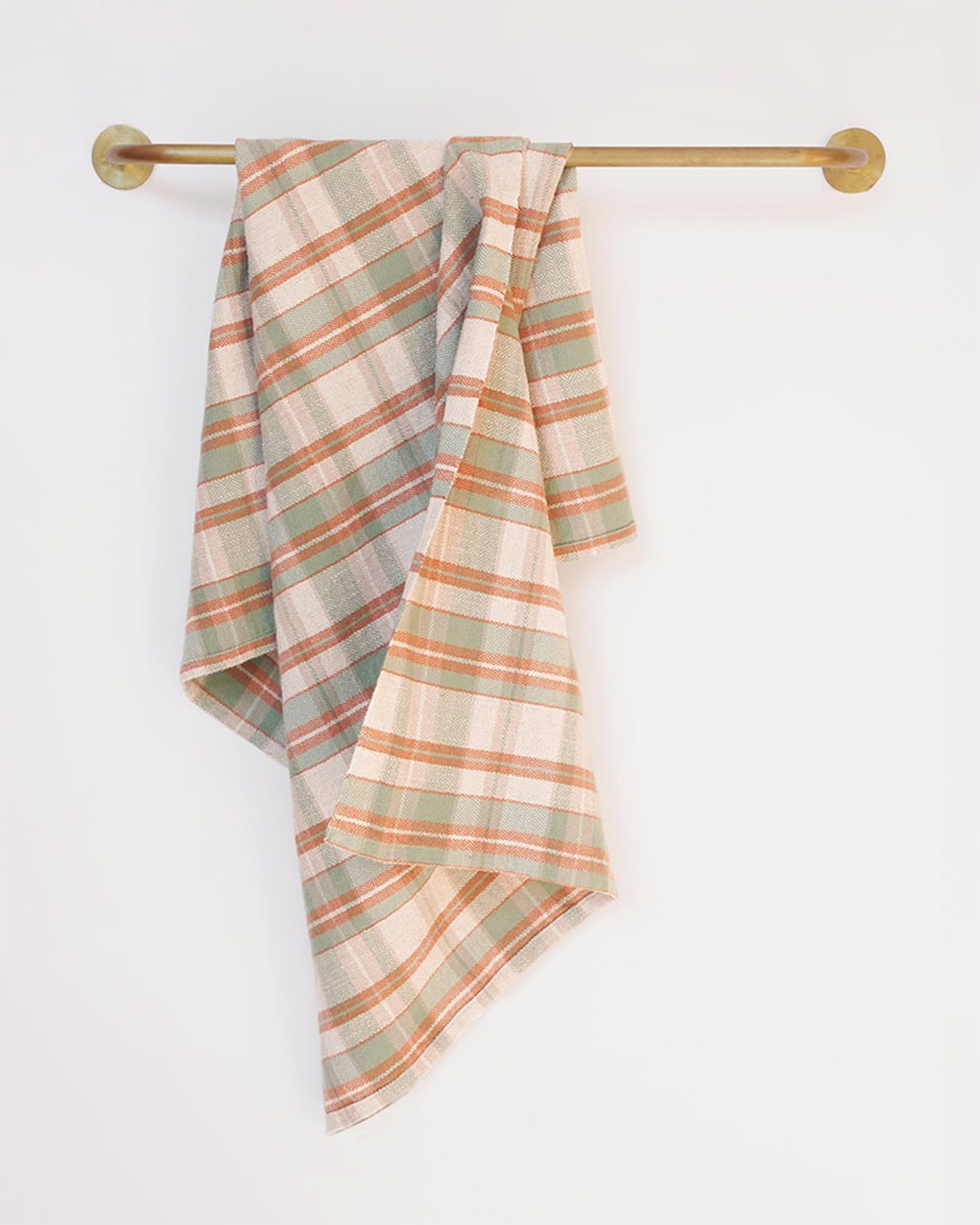 The Faire Cook's Cloth, a soft cotton-linen blend kitchen towel in muted green, orange, and beige plaid, hangs neatly over a brass towel bar on a plain white wall, combining style with durability.