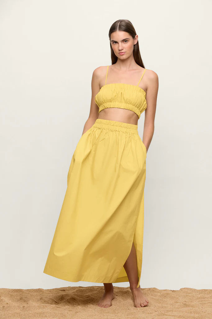 A woman stands barefoot on sand, wearing a yellow two-piece outfit composed of a cropped tank top with thin straps and the Delia Maxi Skirt from Mikoh, featuring high slits. The background is plain off-white. She has long, straight hair and her hands are in her skirt pockets.