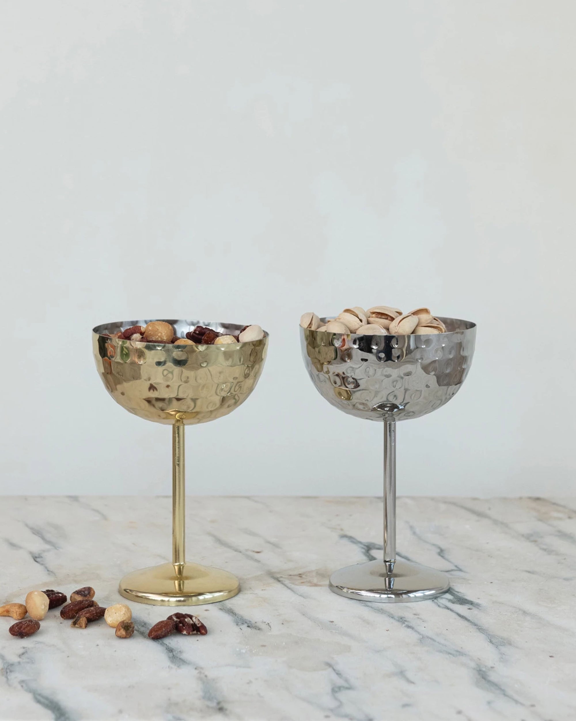 Two Creative Co-op Stainless Steel Coupe Glasses sit on marble—one gold holding mixed nuts and one silver with pistachios. Scattered nuts add to the elegant scene.