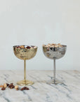 Two Creative Co-op Stainless Steel Coupe Glasses sit on marble—one gold holding mixed nuts and one silver with pistachios. Scattered nuts add to the elegant scene.