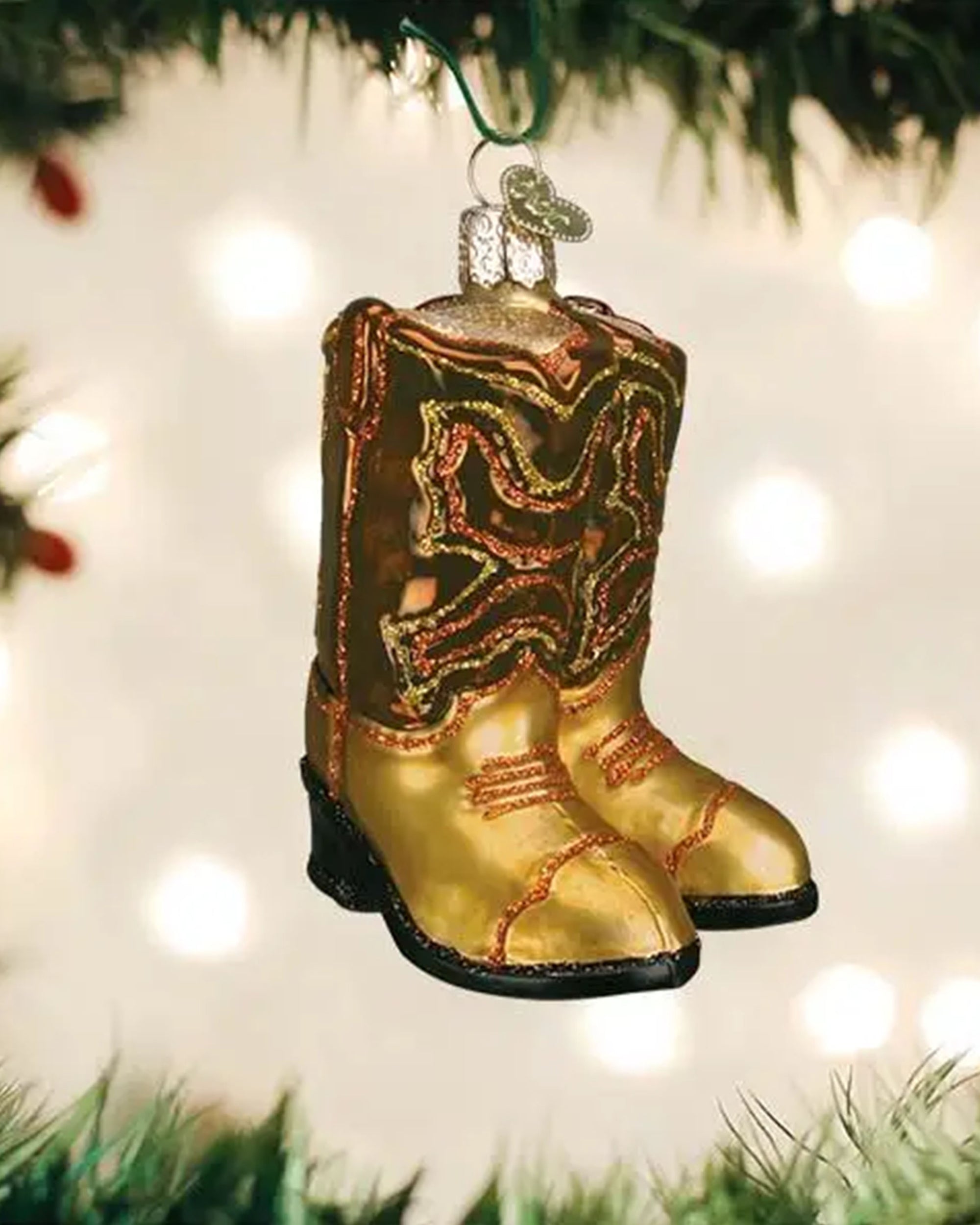 The Faire Old World Ornaments gold cowboy boots, with red and black accents, hang among green branches, adding unique charm to your holiday décor while blurred lights twinkle in the background.