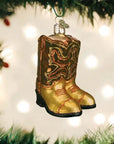 The Faire Old World Ornaments gold cowboy boots, with red and black accents, hang among green branches, adding unique charm to your holiday décor while blurred lights twinkle in the background.