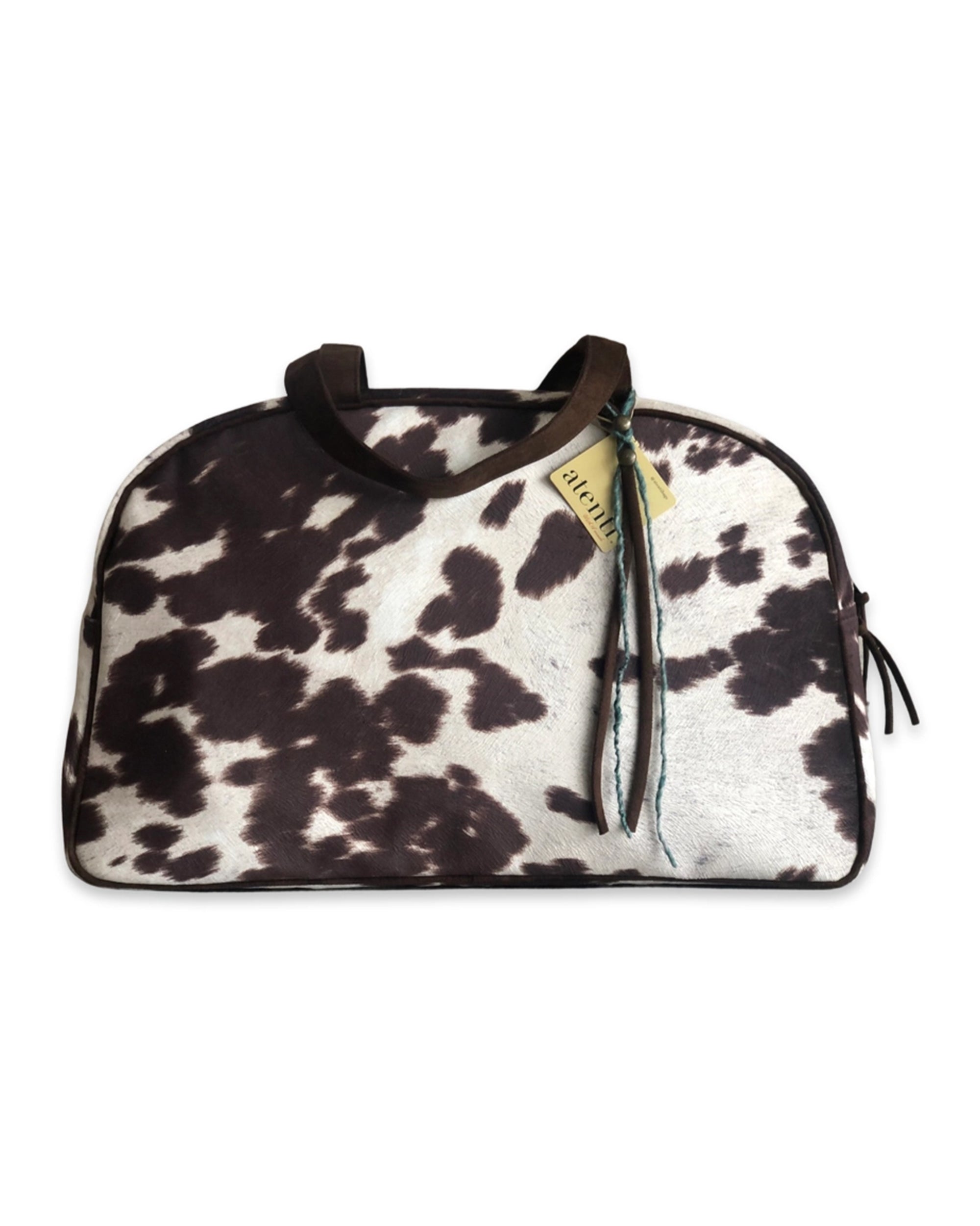 The Faire Oakley Overnighter - Faux Cowhide is a vegan weekender bag featuring a black and white cowhide pattern, brown handles, and a yellow tag, shown on a white background.