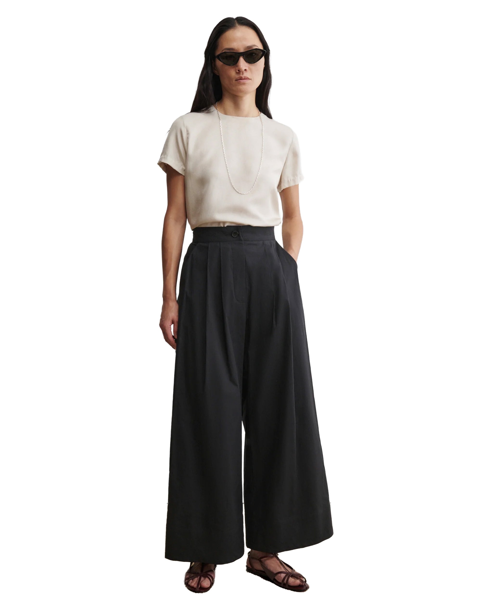 A person stands against a white background with hands in pockets, wearing Rachel Comey’s black Coxsone Pant (organic cotton, wide leg), a beige short-sleeve top, dark sunglasses, a necklace, and sandals.