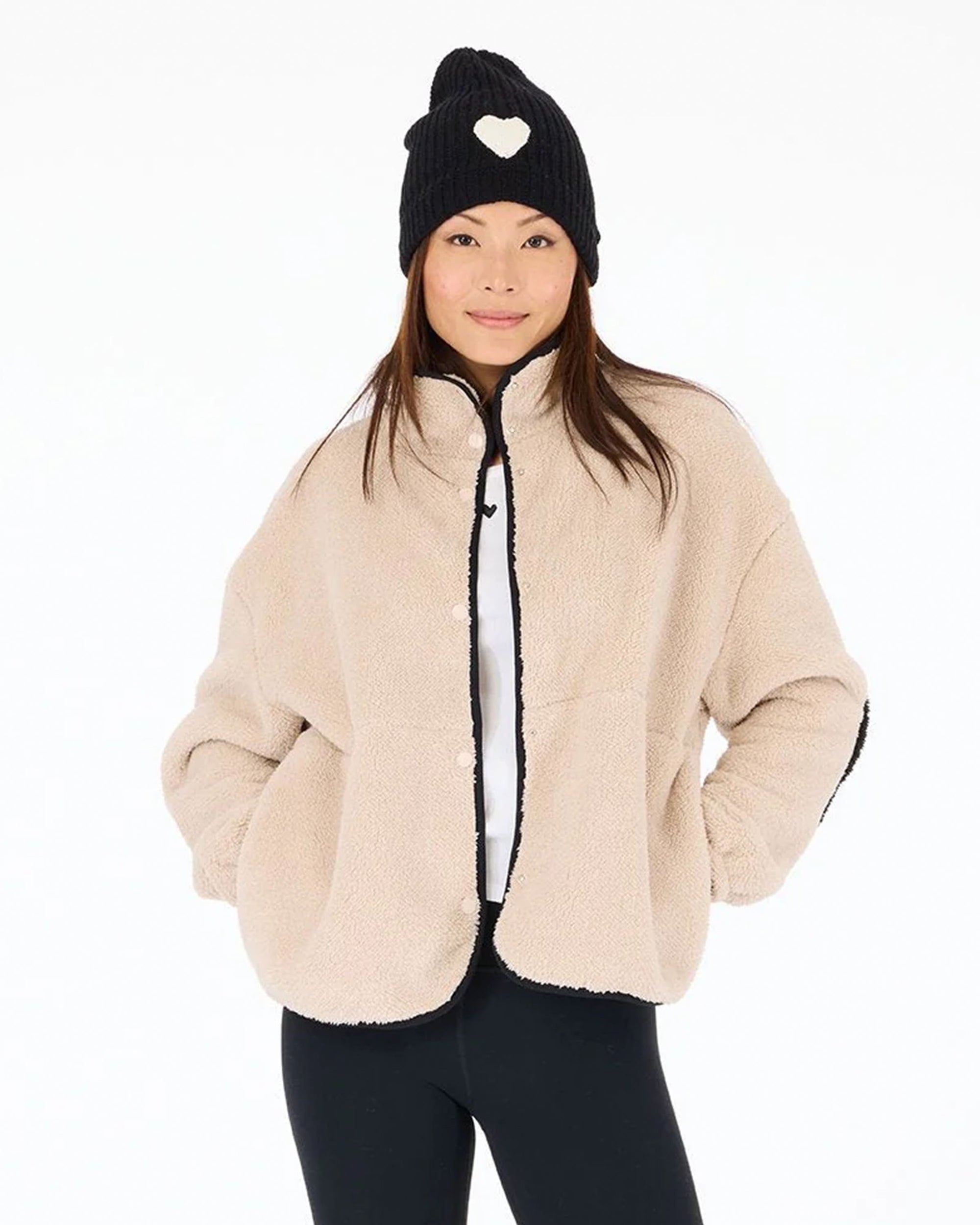 A woman wears a beige Kerri Rosenthal Coze Magoze jacket with heart elbows, black leggings, and a black knit beanie with a white heart, smiling with hands in pockets against a plain white background.