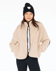 A woman wears a beige Kerri Rosenthal Coze Magoze jacket with heart elbows, black leggings, and a black knit beanie with a white heart, smiling with hands in pockets against a plain white background.