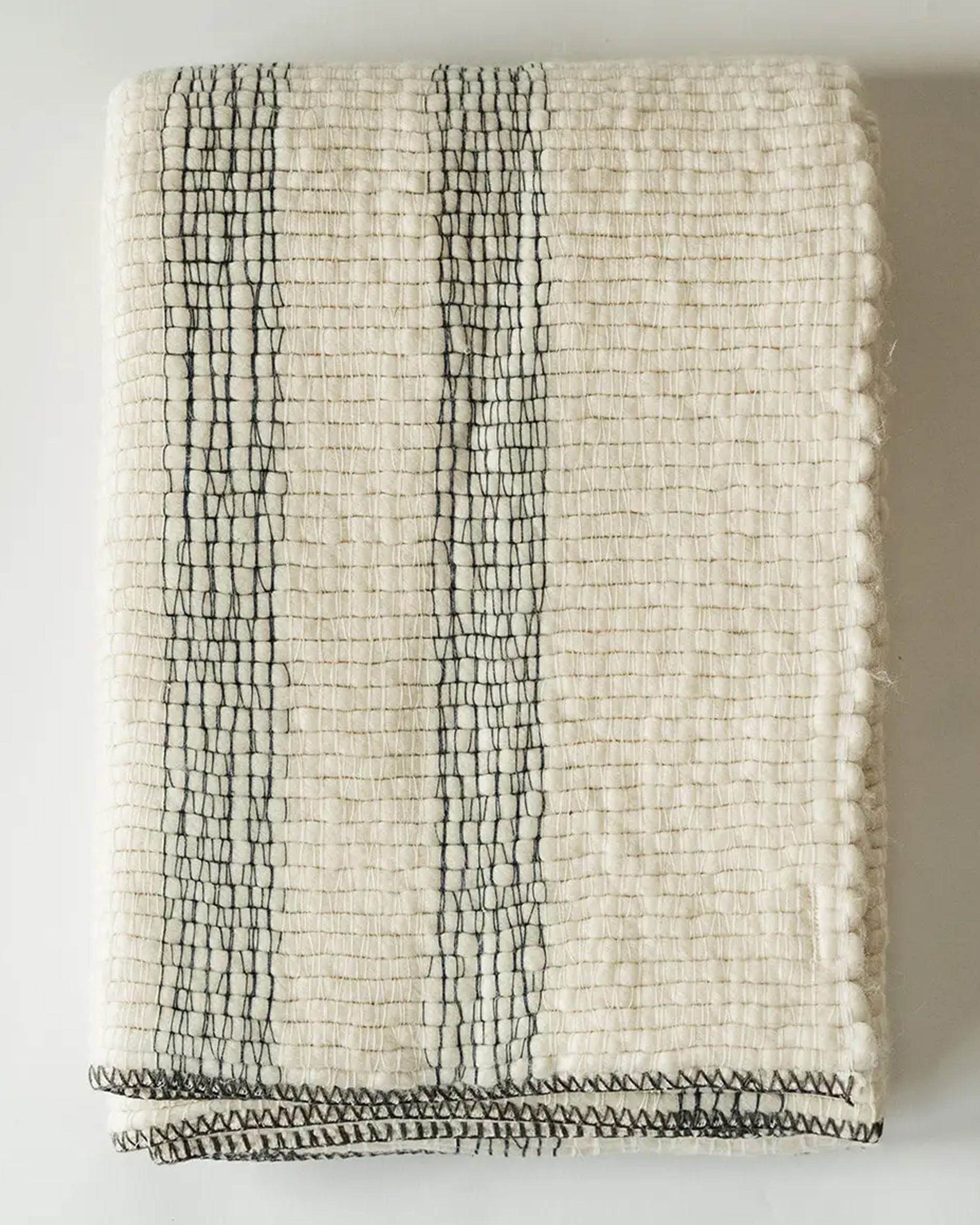 The Faire Cozy Stripe Throw, a cream-colored wool-blend with black vertical stripes and stitched edges, is folded neatly on a white surface.