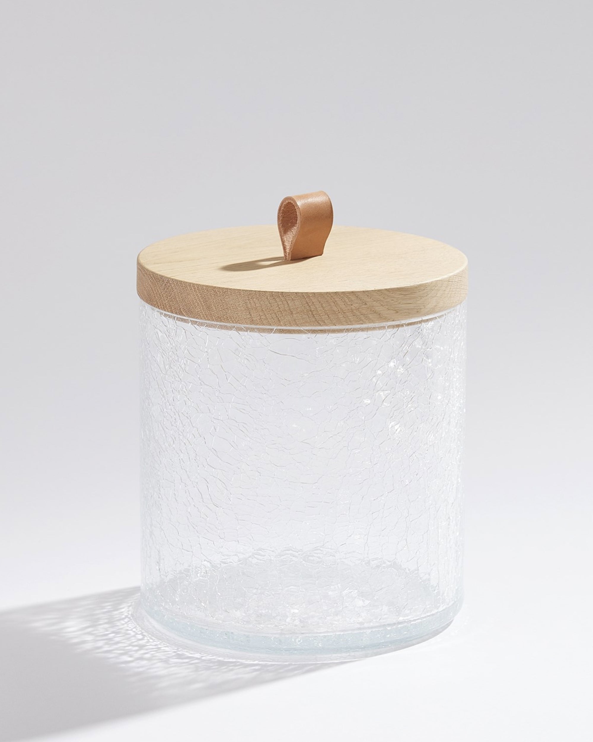 The BB-1.10080 Crackle Glass Canister MD by Global Views features handcrafted Portuguese crackle glass with a textured finish, finished with a smooth oak lid and brown leather loop handle, displayed elegantly against a plain background.
