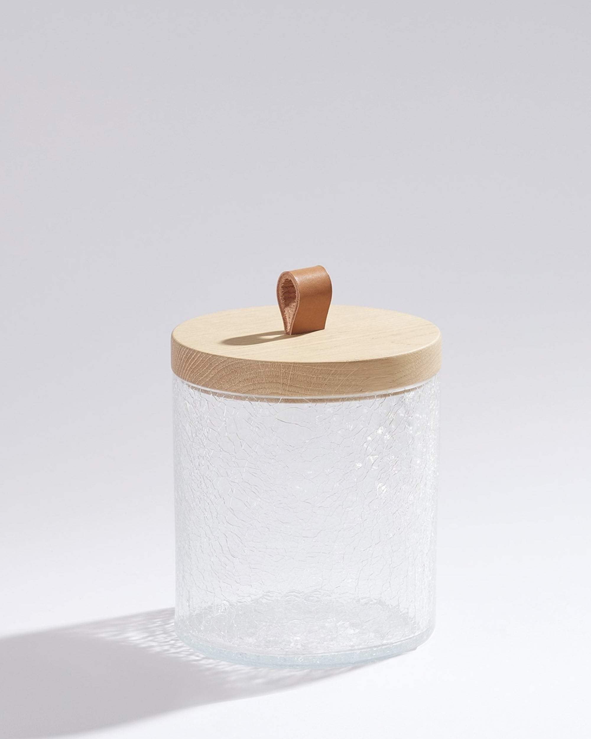 The BB-1.10081 Crackle Glass Canister SM by Global Views features clear, textured glass with a light oak lid and brown leather loop handle, casting a soft shadow—ideal for handcrafted storage in any setting.