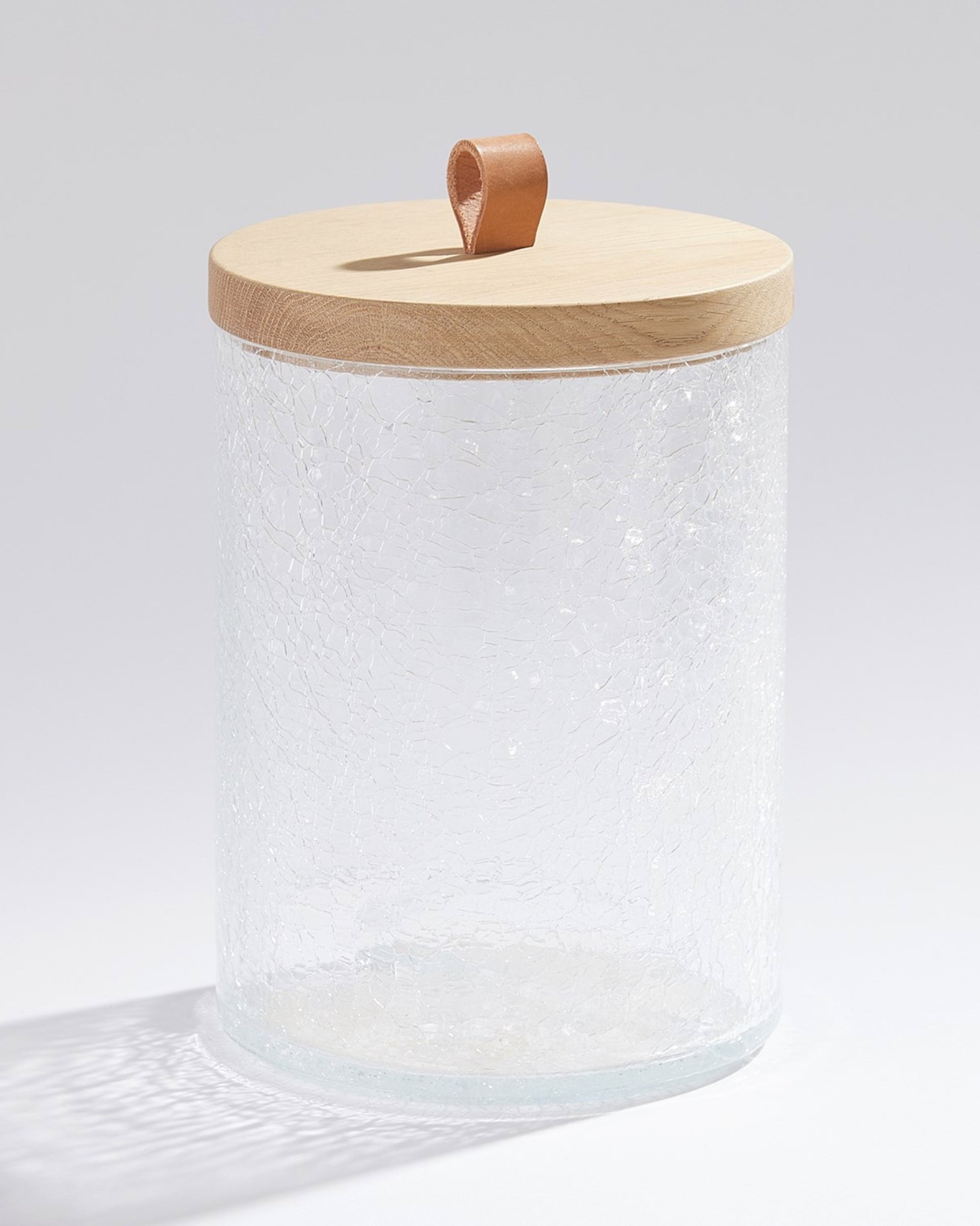 The Global Views BB-1.10079 Crackle Glass Canister LG is a clear, cylindrical jar with a unique crackled texture, topped by a smooth oak lid and tan leather loop handle. Stylishly empty, it sits on a white surface, ideal for chic home decor displays.