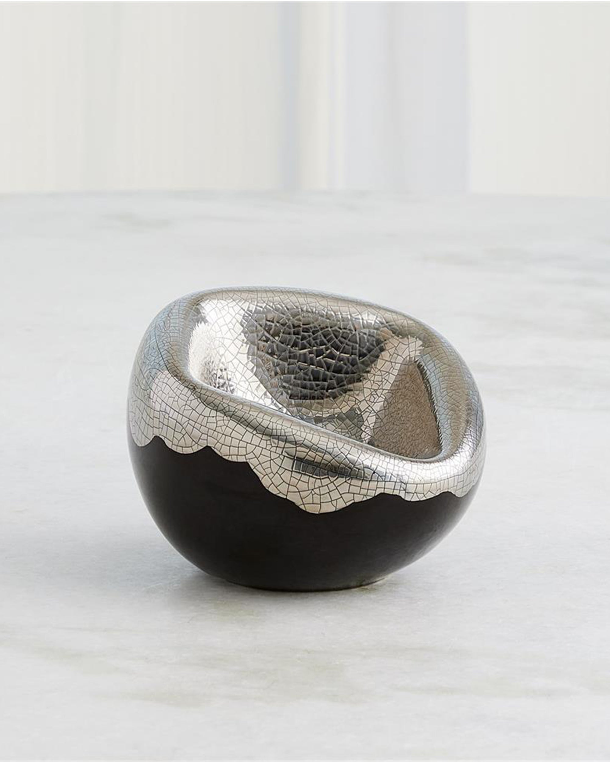 The Global Views 3.31835 Squished Bowl Platinum Crackle is a small, round, black Italian ceramic bowl with an irregular metallic silver rim and a platinum crackle interior, shown on a light-colored surface with a blurred background.