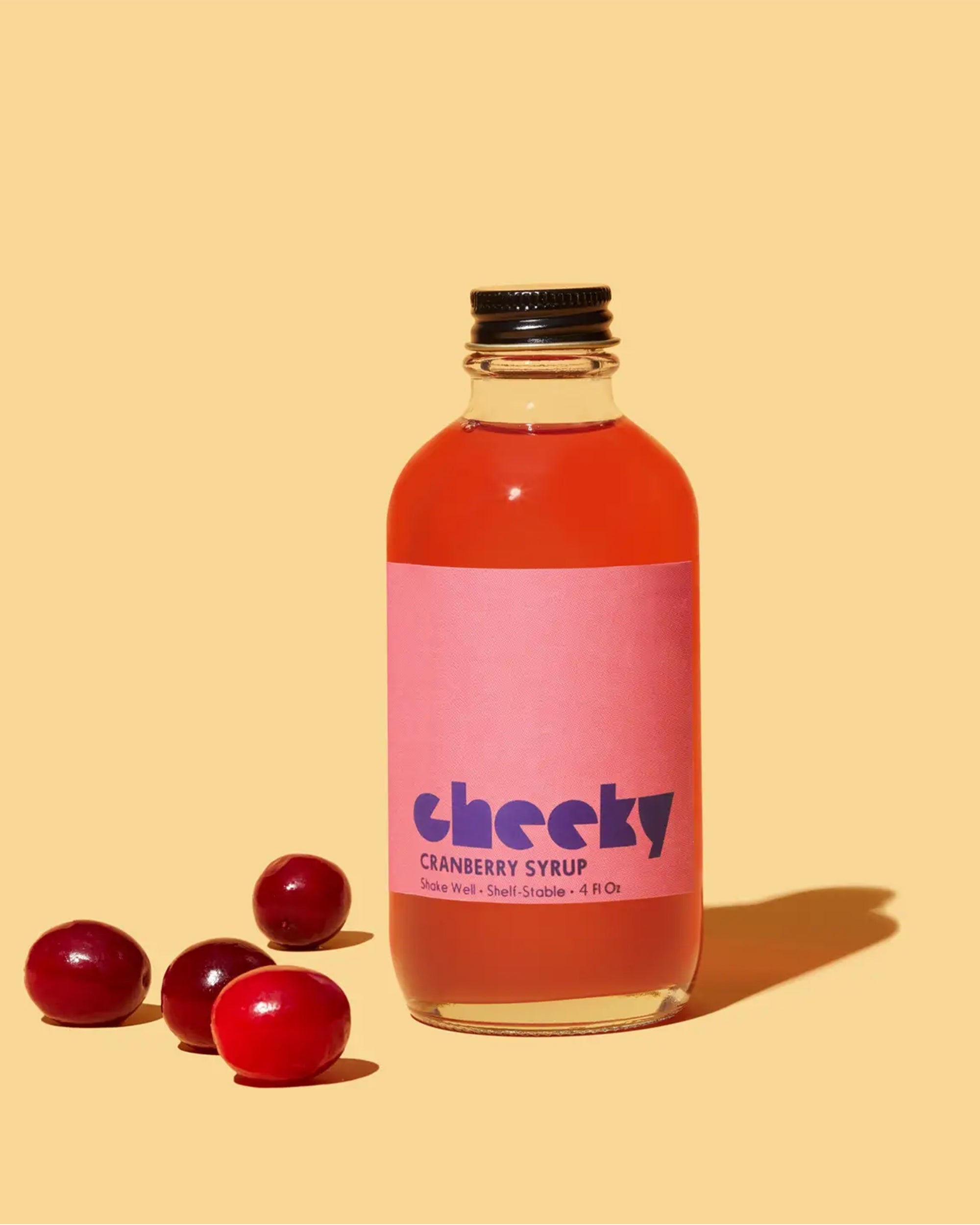 A clear glass bottle of Faire Cranberry Syrup 4oz with a pink label rests on a beige surface beside four cranberries—ideal for festive drinks or recipes. The background is a solid light beige.