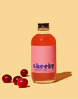 A clear glass bottle of Faire Cranberry Syrup 4oz with a pink label rests on a beige surface beside four cranberries—ideal for festive drinks or recipes. The background is a solid light beige.
