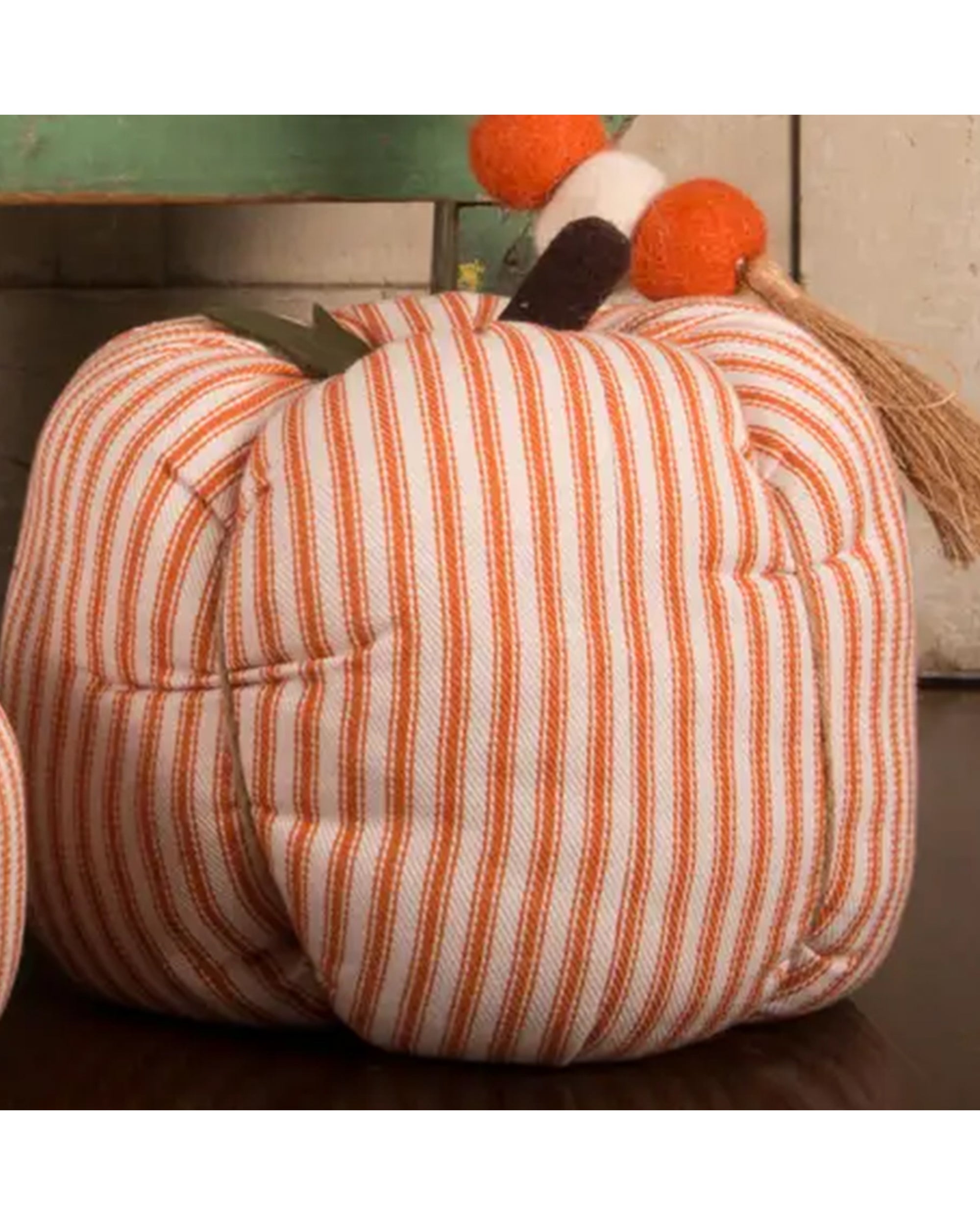 The Faire Pumpkin is a soft fabric ornament with orange and white stripes, a decorative stem featuring orange pom-poms and beige tassels—perfect for cozy farmhouse decor.