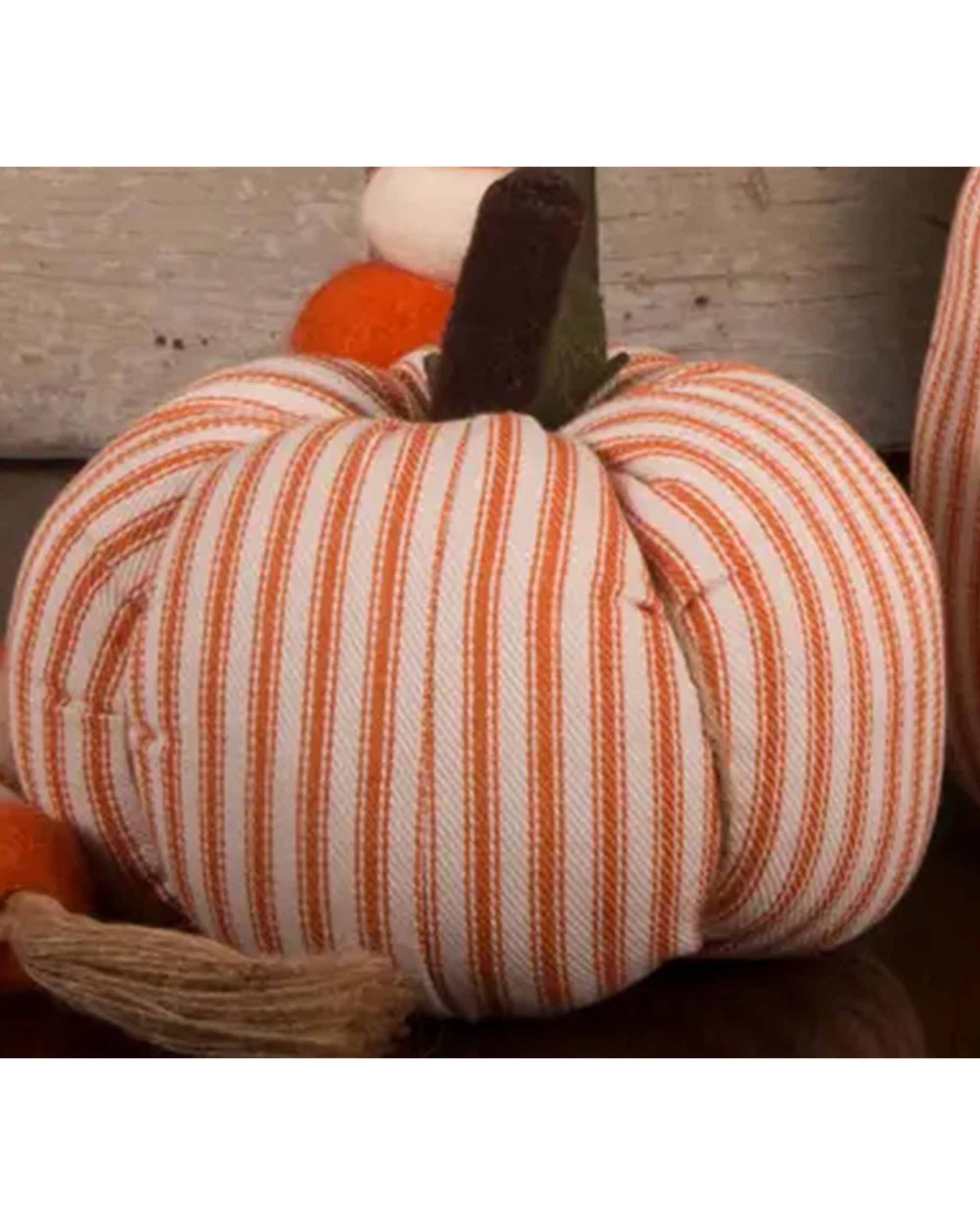 The Faire Pumpkin, featuring soft orange and white vertical stripes with a brown fabric stem, sits on a dark surface for charming farmhouse decor; another plaid pumpkin appears in the background.