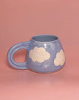 The Cute Mug by Faire is a handmade pottery mug with a rounded shape, large handle, and white cloud patterns, displayed on a pink background.