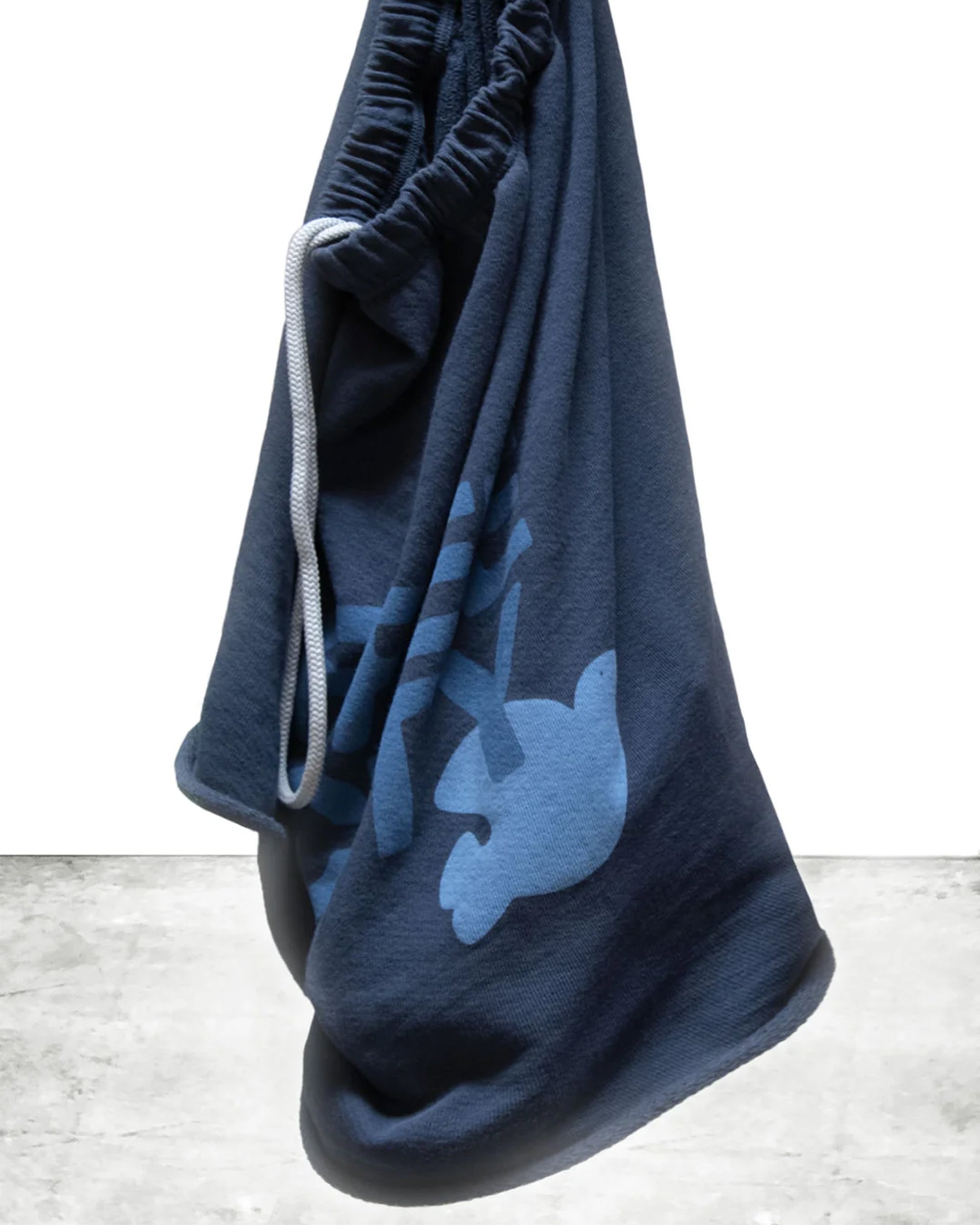 The FreeCity Cutoffs bag by Free City (sparrow, LLC) is a dark blue French terry drawstring with a light blue bird and branch design, displayed against a white background and gray concrete floor.
