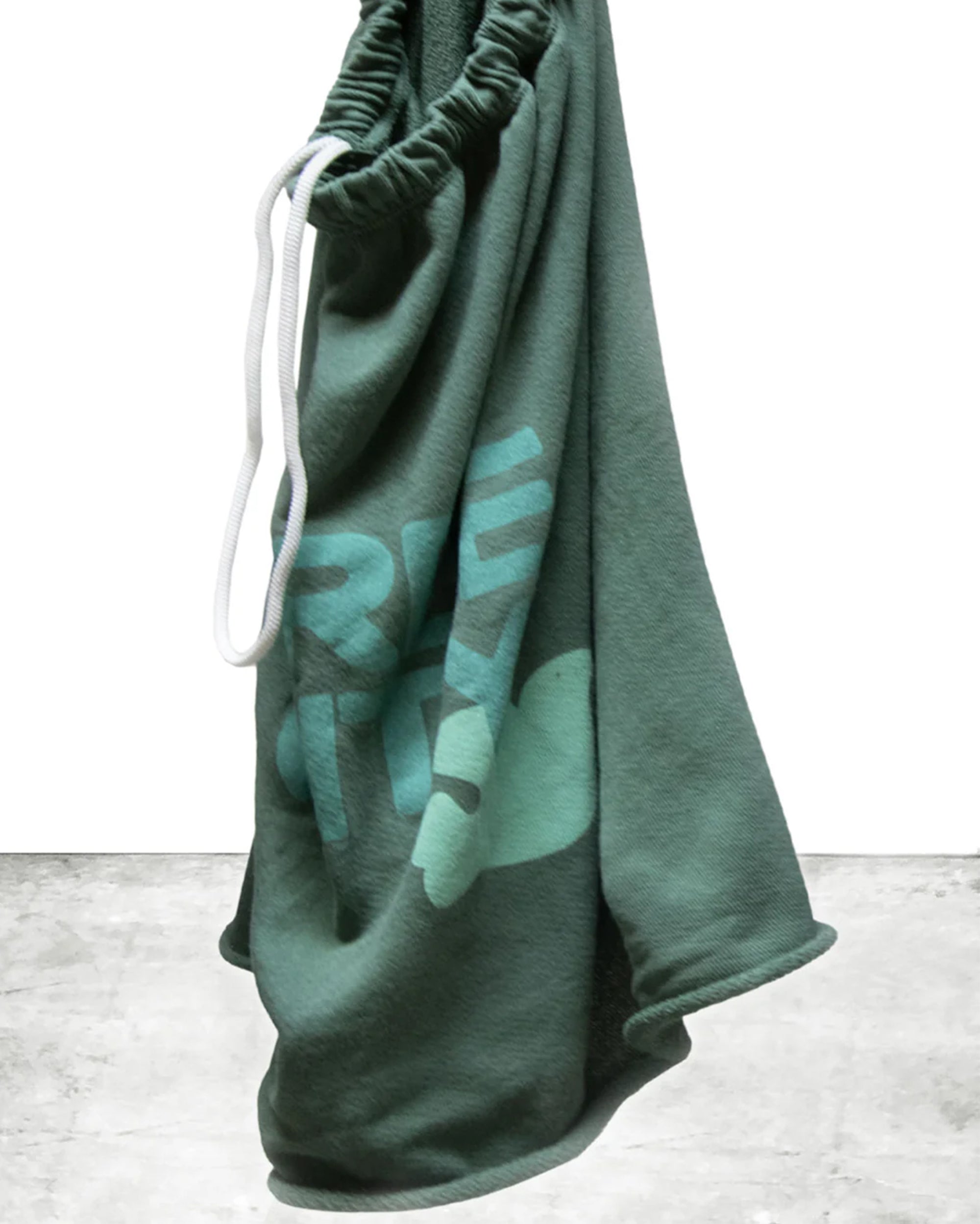 A pair of green FreeCity Cutoffs by Free City (sparrow, LLC) with white drawstrings hangs against a white background and gray floor, showing a large light blue graphic on the leg. Made from lightweight French terry.