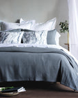 A bed features the Eastern Accents Tegan Matelasse Coverlet in textured gray 100% cotton with a mitered hem, white sheets, marbled black-and-white pillows, and a plant on a small wooden table by sheer white curtains.