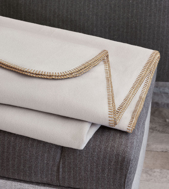 The Pattinson Blanket Stitch Coverlet by Eastern Accents, in light beige flannel with brown stitched edges, is folded atop a dark gray pinstriped blanket against a textured Carrara Fog background.