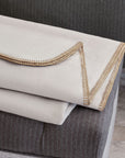 The Pattinson Blanket Stitch Coverlet by Eastern Accents, in light beige flannel with brown stitched edges, is folded atop a dark gray pinstriped blanket against a textured Carrara Fog background.