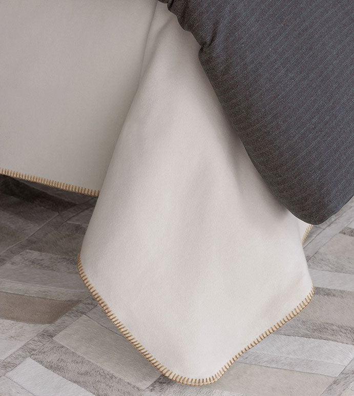 A close-up of a bed corner with the Eastern Accents Pattinson Blanket Stitch Coverlet in cream, featuring tan blanket stitch edging. A dark gray striped comforter is partly on top, and the floor shows Carrara Fog patchwork tiles.