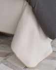 A close-up of a bed corner with the Eastern Accents Pattinson Blanket Stitch Coverlet in cream, featuring tan blanket stitch edging. A dark gray striped comforter is partly on top, and the floor shows Carrara Fog patchwork tiles.
