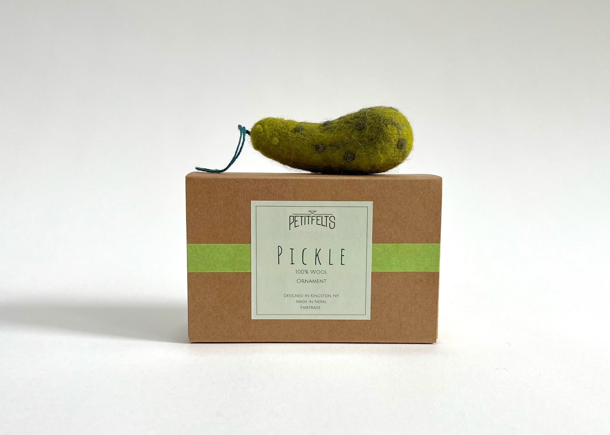 A small Felted Ornament by Faire, shaped like a pickle with a green string stem, sits atop a brown box labeled "Pettifelts Pickle" with a green stripe, set against a plain white background.