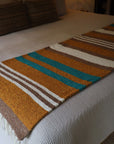 Handwoven striped blanket in brown, gold, teal, and white on bed; Bungalow Furniture & Accessories.