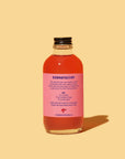 A clear glass bottle with pink liquid and a pink "Cranberry Syrup 4oz" label from Faire sits on a solid light peach background. The label lists ingredients and instructions for preparing this festive drink.