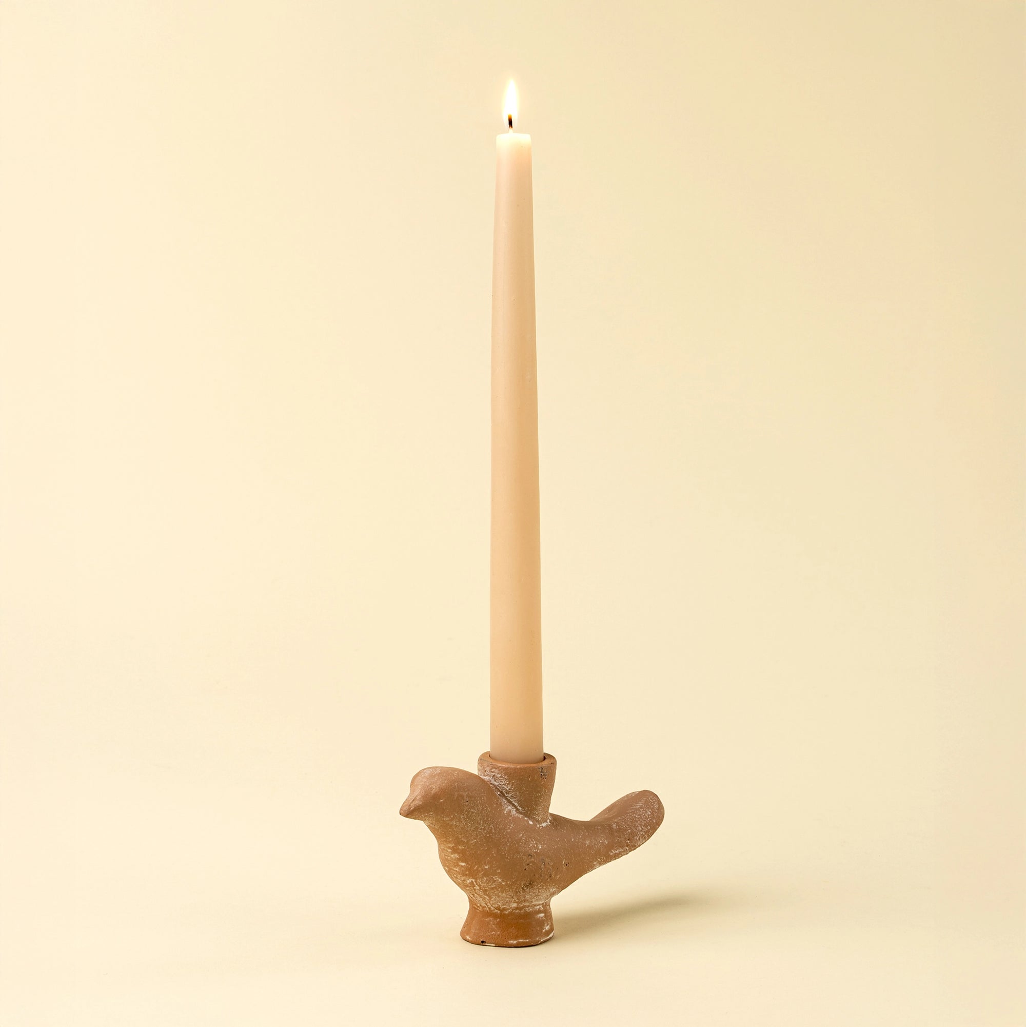 A lit beige taper candle sits in the sculptural Indaba Wren Candle Holder, bird-shaped and set against a light cream background—an ideal rustic accent for your decor.