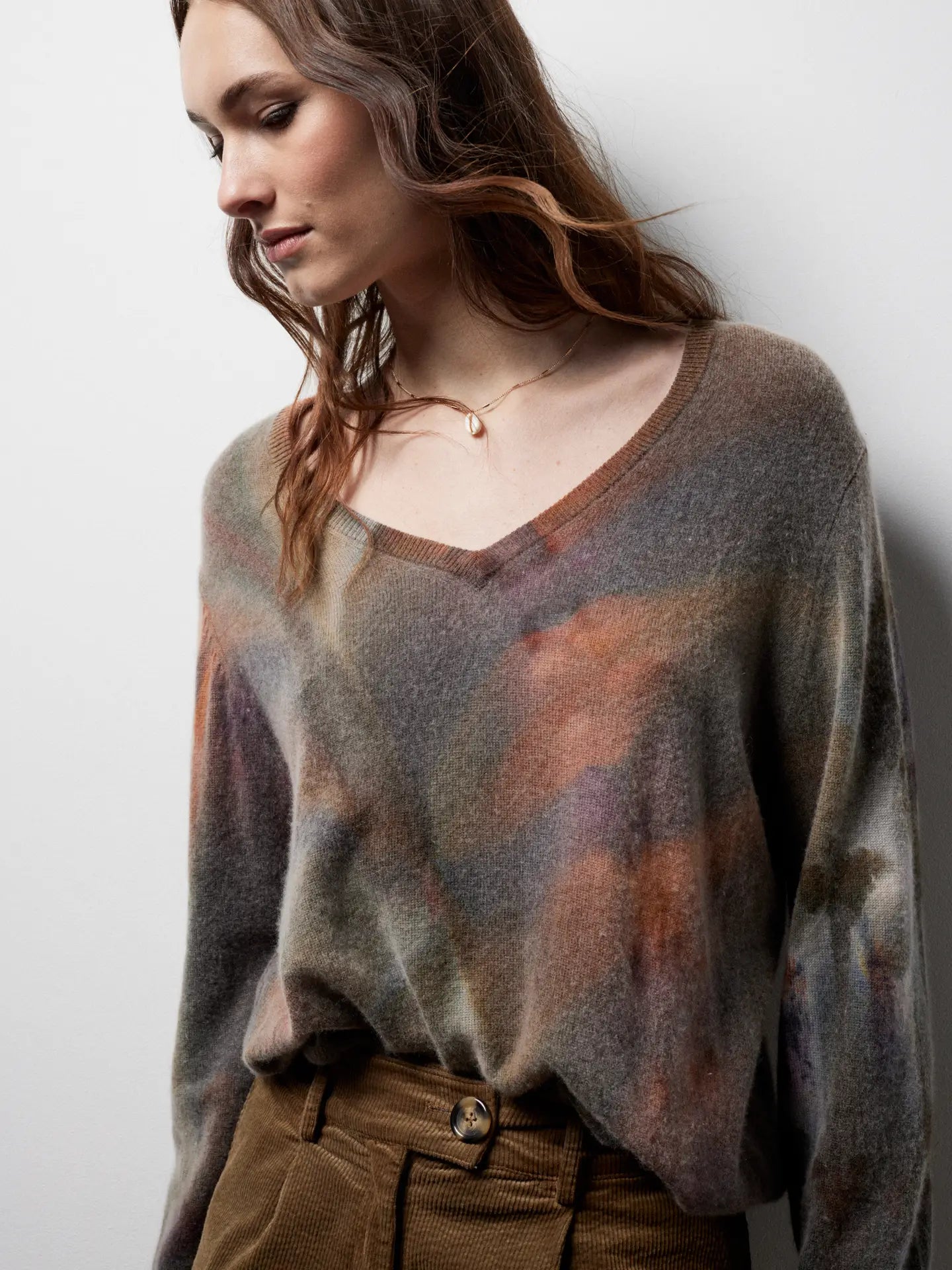 A woman with wavy brown hair looks down while wearing the Riverside Tool & Dye Cashmere V-neck sweater, featuring a blurred multicolor pattern, paired with brown corduroy pants against a plain white background.