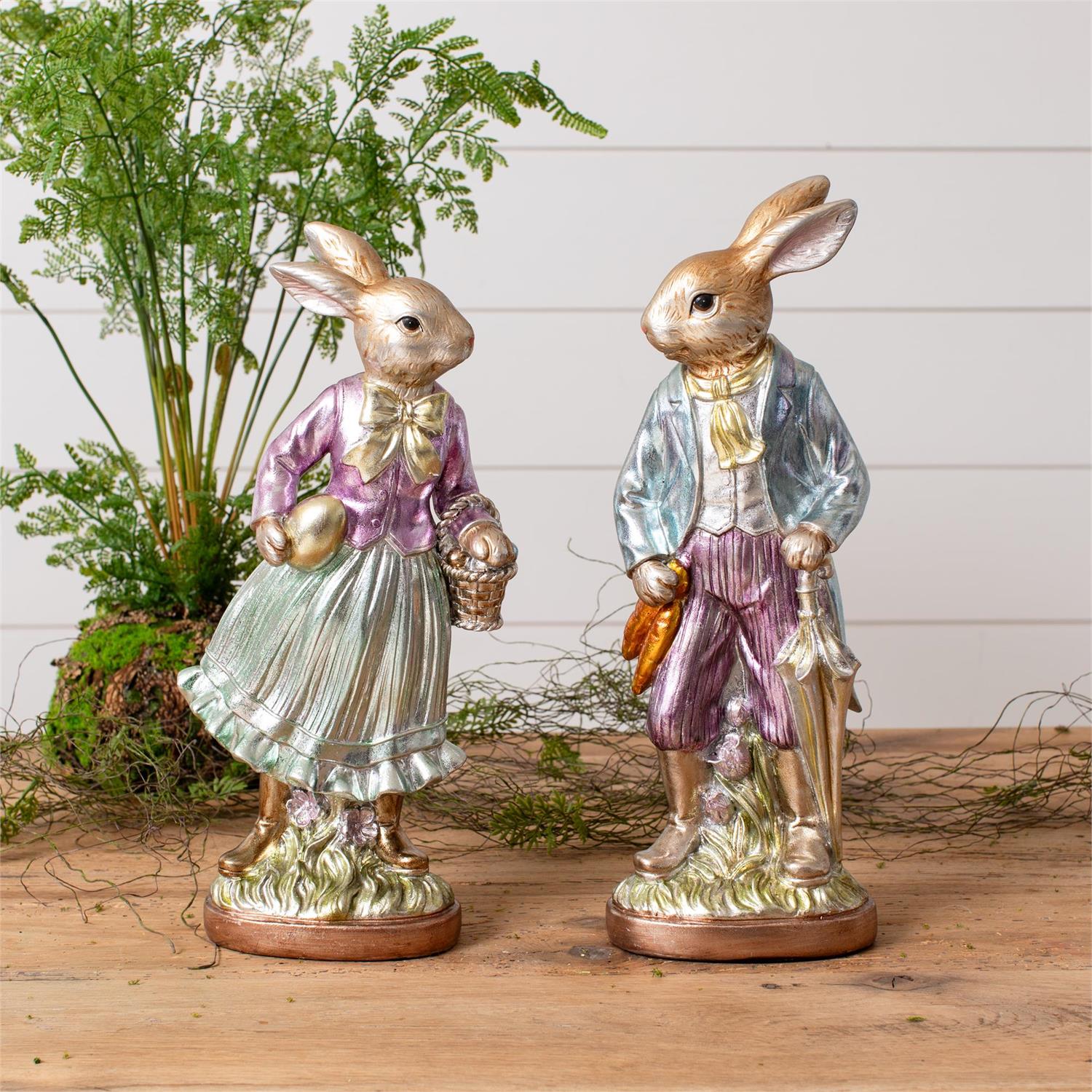 The Faire Vintage Foil Rabbit figurines, dressed in vintage-style clothing, stand together on a wooden surface with greenery—perfect heirloom-inspired decor for charming Easter displays.