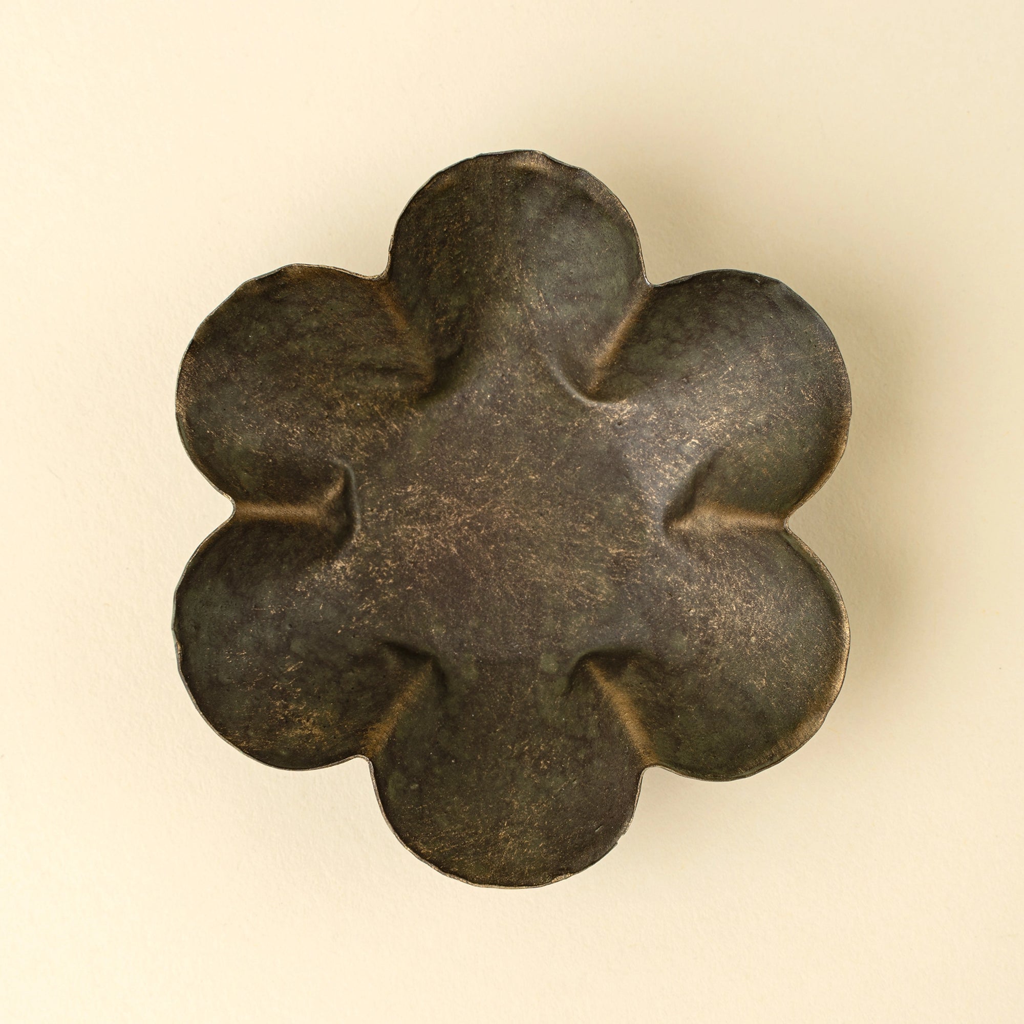 The Faire Florentine Catchall Dish, Daisy, features a dark metallic six-petaled flower design with textured, gently grooved petals and a vintage finish, displayed against a light beige background.