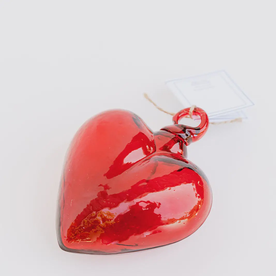 The Glass Heart Medium by Faire is a handcrafted, glossy, red glass heart-shaped ornament that evokes Mexican tradition. It rests on a white surface and features a string with a white tag.