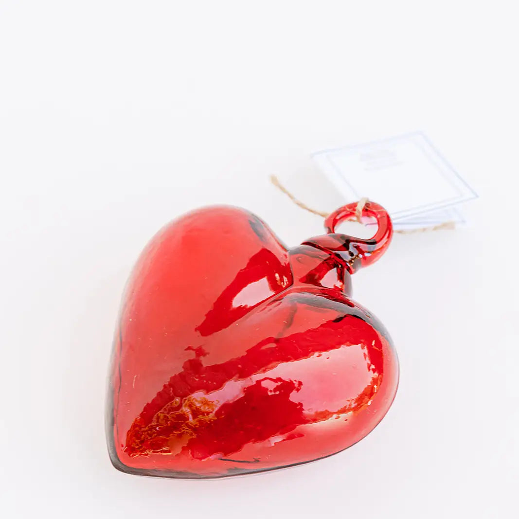 The Faire Glass Heart Small ornament is a glossy red blown glass piece with a top loop and an attached tag, reminiscent of rich Mexican tradition. This handcrafted ornament is beautifully reflective and elegantly displayed against a plain white background.