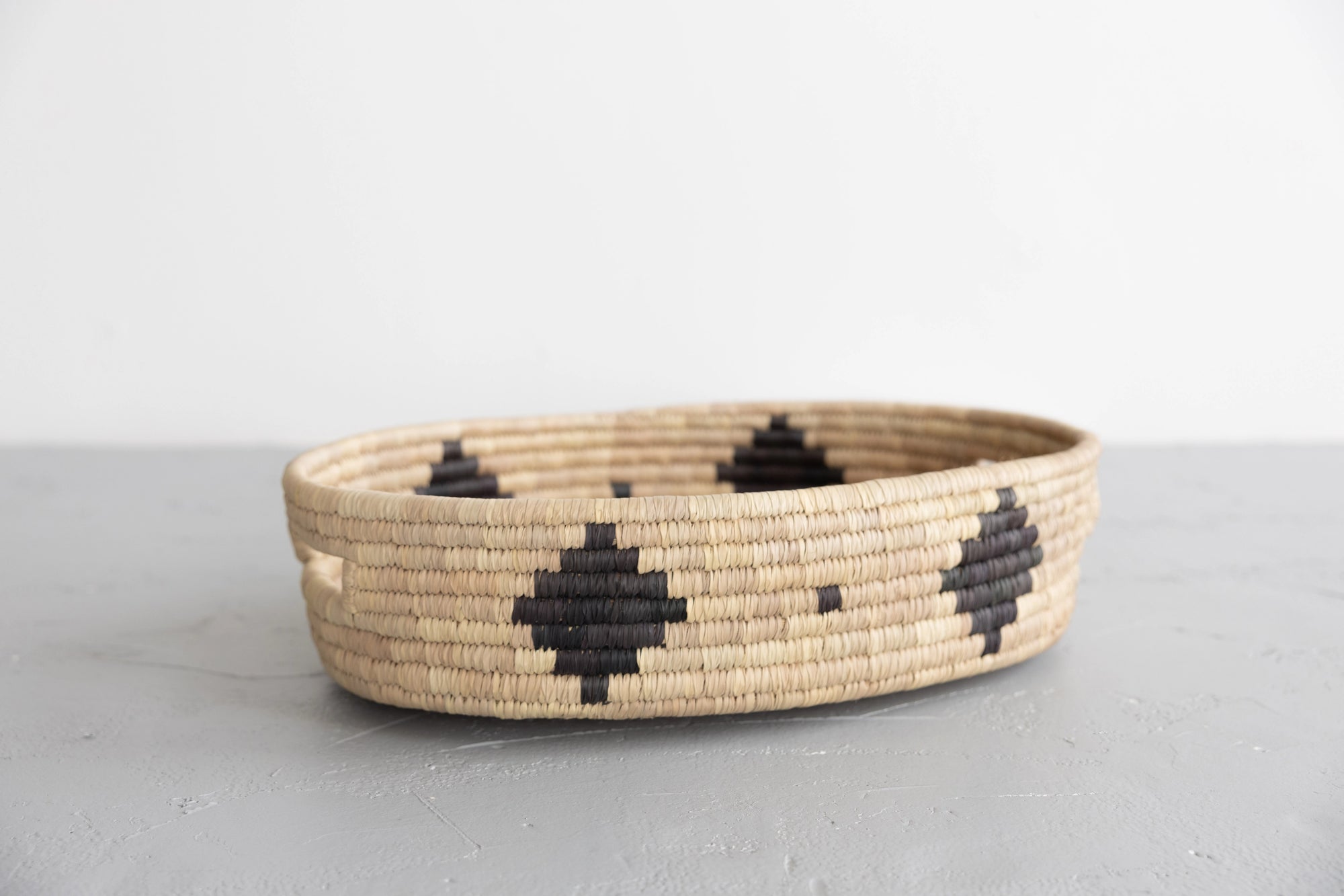 The Faire Palm Leaf Oval Basket, a handwoven beige tray with black geometric patterns crafted by fair trade artisans, rests on a gray surface against a white background.