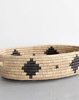 The Faire Palm Leaf Oval Basket, a handwoven beige tray with black geometric patterns crafted by fair trade artisans, rests on a gray surface against a white background.