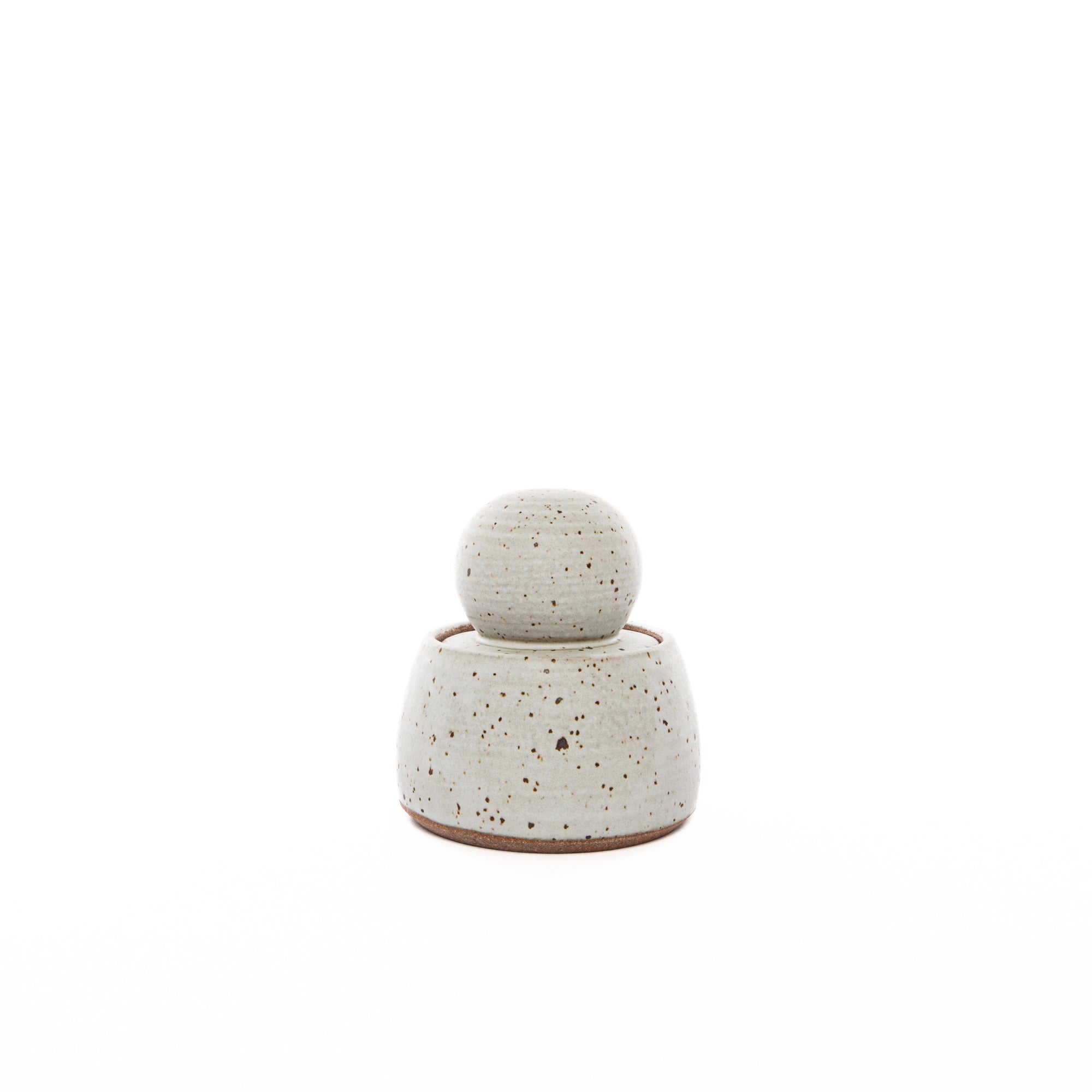 The Alabaster White Stash Jar by Faire, a small round stoneware jar with a matching lid in light beige and dark brown flecks, finished with a matte glaze, is centered on a white background.