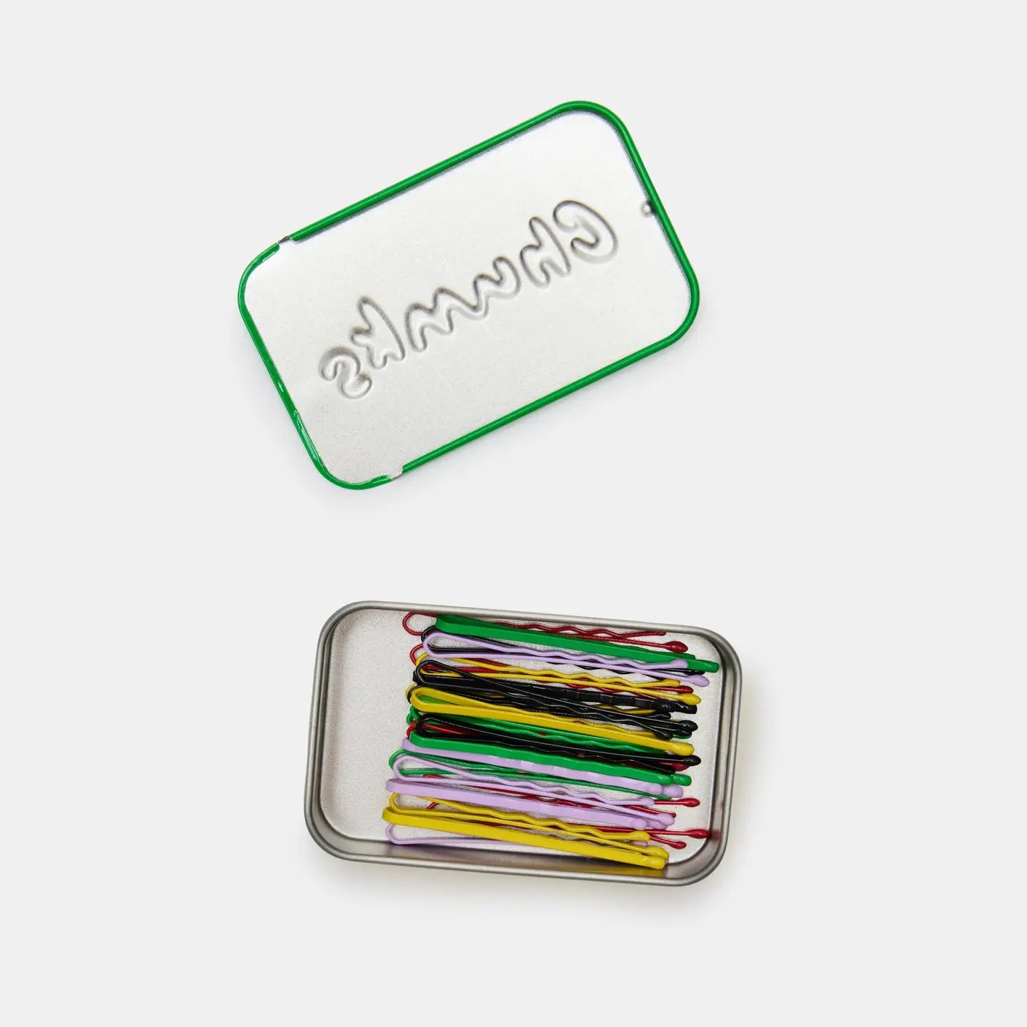 A compact rectangular metal tin with a green-edged lid labeled "CHUNKS" by Faire holds neatly arranged multicolored bobby pins. The open tin and its lid are displayed on a plain white background.