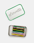 A compact rectangular metal tin with a green-edged lid labeled "CHUNKS" by Faire holds neatly arranged multicolored bobby pins. The open tin and its lid are displayed on a plain white background.