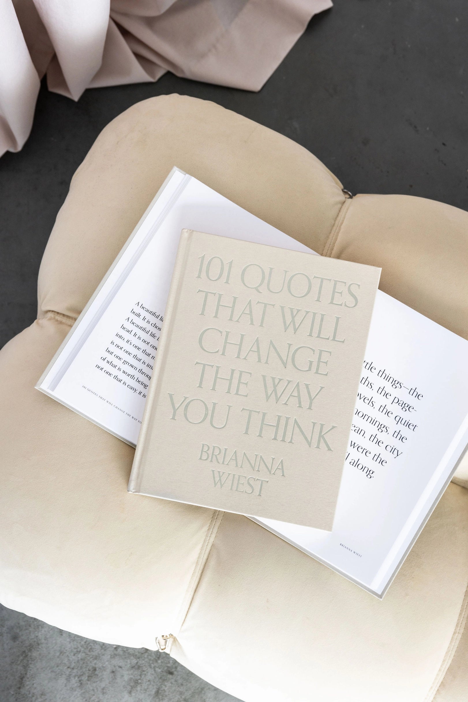 The beige "101 Quotes That Will Change the Way You Think" by Faire rests atop an open book on a light cushioned seat, providing daily inspiration with selected quotes.