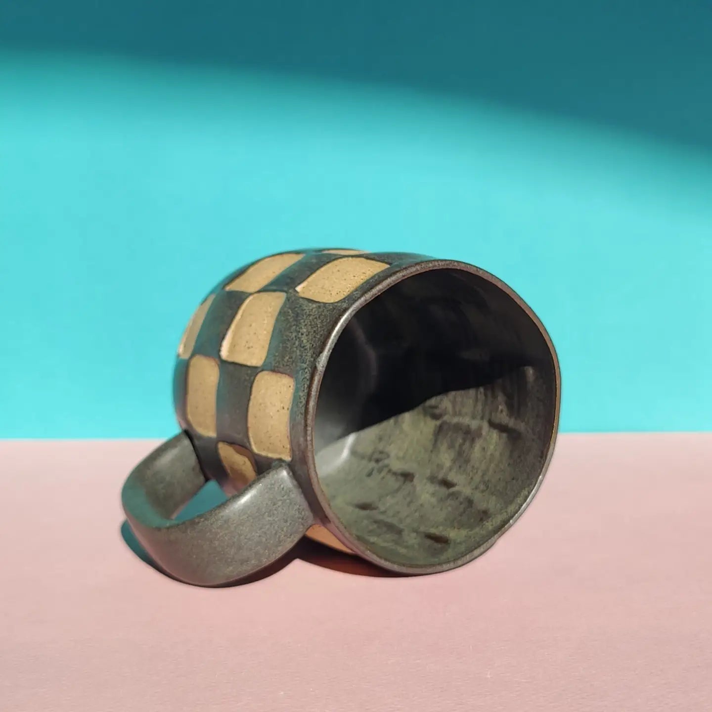 The OC Checkerboard Mug by Faire, featuring a beige and green checkered stoneware design, rests on its side on a pink surface with a blue backdrop and shadow.
