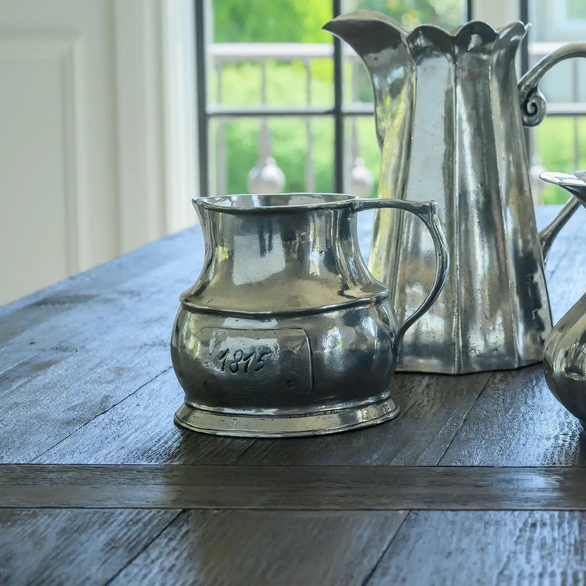 The Vintage 1815 Large Pitcher by Faire, crafted in Italy, sits on a dark wooden table near a window, with two other metal pitchers behind it as bright natural light streams in.